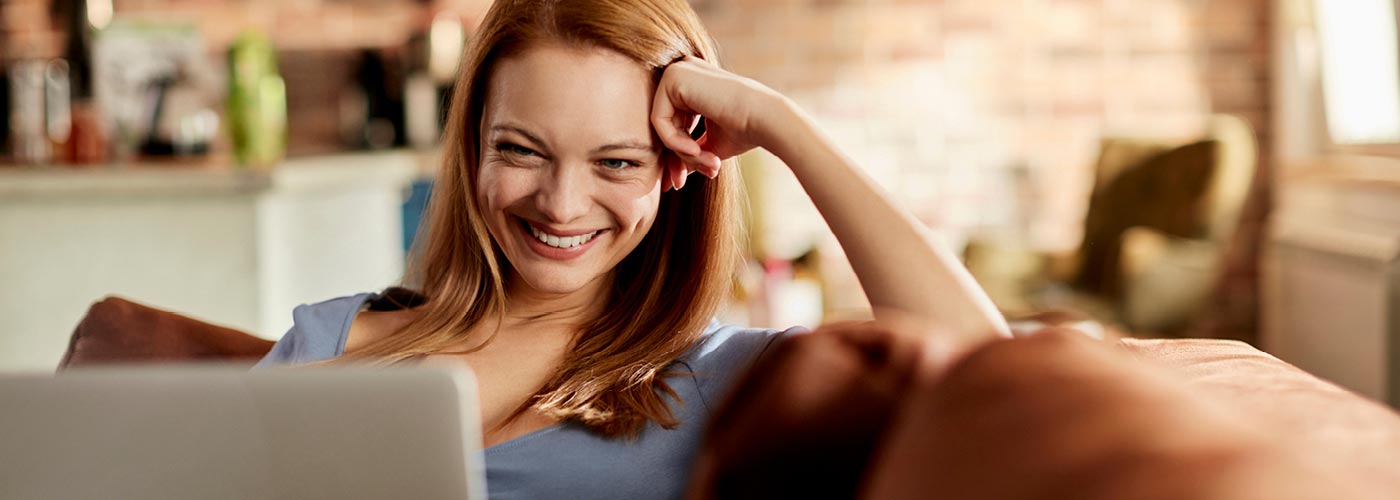 Eine Frau sitzt mit ihrem Tablet auf einer Couch, die Küche im Hintergrund.
